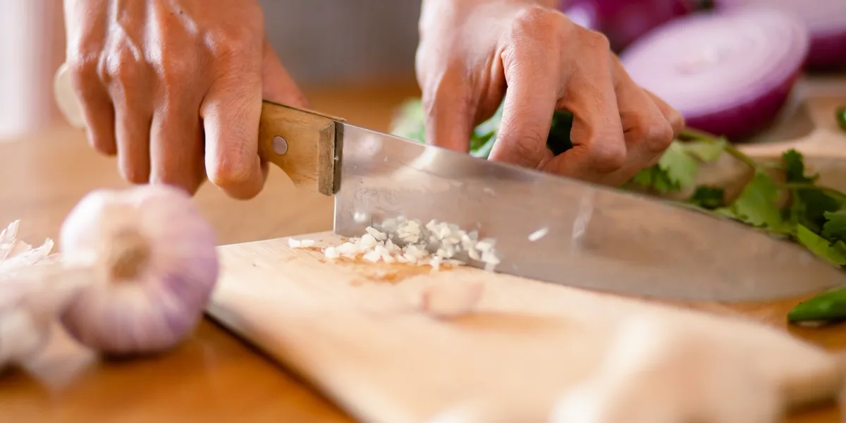 Hands mincing garlic with a chef's knife on a wooden cutting board, with red onion and herbs behind
